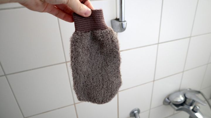 Ein Peelinghandschuh wird von einer Hand gehalten, vor wei&szlig;en Badezimmerfliesen nahe einem Wasserhahn in einer Dusche.