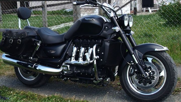 Eine gl&auml;nzende schwarze Motorrad steht auf einer Wiese, umgeben von einem Zaun und einem Geb&auml;ude im Hintergrund.