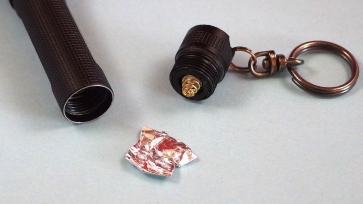 Eine zerknitterte Alufolie liegt neben einem Taschenlampenkopf und einem Schl&uuml;sselring auf einem hellen Untergrund.