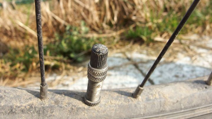 Ein Fahrradventil steht im Fokus, w&auml;hrend ein Luftverlust im Reifen sichtbar wird, umgeben von gr&uuml;nem Gras und Erde.