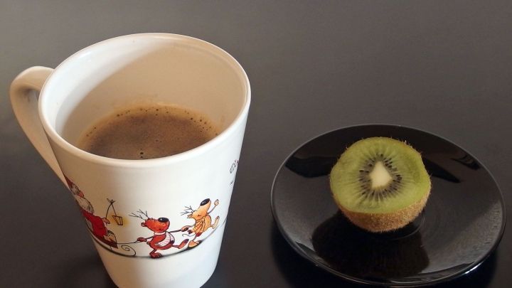 Eine Tasse Kaffee steht neben einer Kiwi auf einem schwarzen Teller, auf einem glatten Tisch, ideal f&uuml;r Kater und Kopfschmerzen.