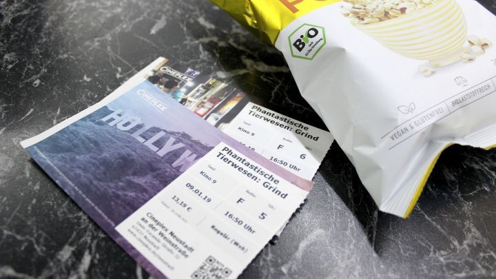 Zwei Kinotickets liegen neben einem ge&ouml;ffneten Beutel Popcorn auf einem Tisch vor einem Filmstart.