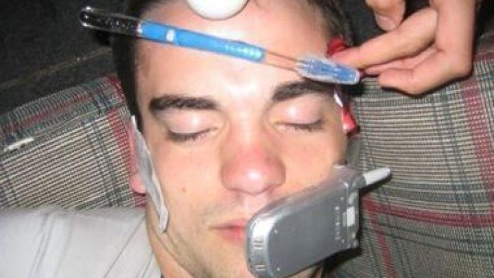 Ein Mann schl&auml;ft mit verschiedenen Gegenst&auml;nden wie einem Handy und einem Tischtennisball auf seinem Gesicht, w&auml;hrend jemand ihm einen Stift an die Stirn h&auml;lt.