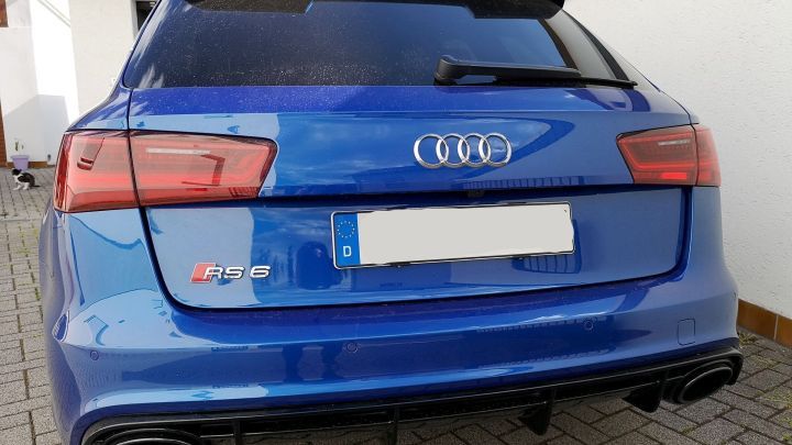 Ein blauer Audi RS6 parkt sauber in einem gepflasterten Hof neben einer wei&szlig;en Wand.