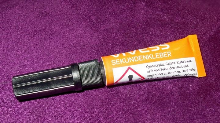 Ein Tubenbeh&auml;lter mit Sekundenkleber liegt auf einem lila Stoff, bereit zum Kleben. Gef&auml;hrliche Chemikalien sind gekennzeichnet.