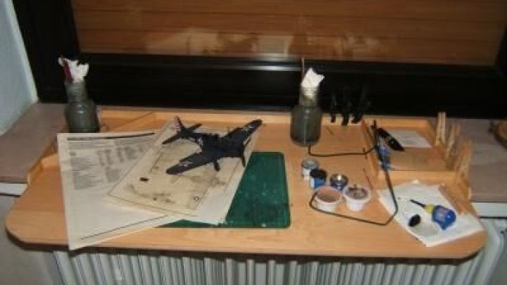 Modellflugzeug auf Bastelbrett aus Laminatresten mit Farben und Pinseln neben Bauanleitung, am Fenster in Bastelumgebung.