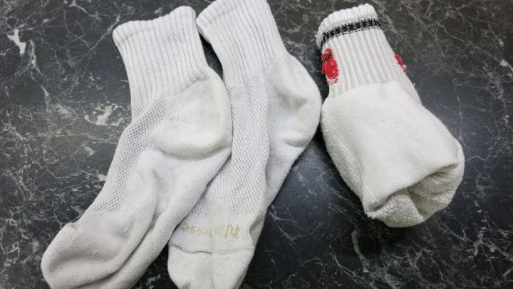 Drei wei&szlig;e Socken liegen chaotisch auf einer dunklen, marmorierten Oberfl&auml;che, zwei sind kurz, eine hat einen bunten Rand.