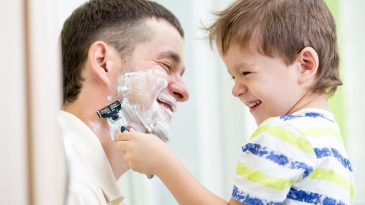 Ein Kind rasiert spielerisch einen Mann mit Rasierschaum im Gesicht im Badezimmer, beide lachen freudig miteinander.