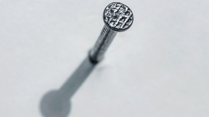 Ein gl&auml;nzender Nagel steht auf einer hellen Oberfl&auml;che, w&auml;hrend sein Schatten darauf zu sehen ist, was die Umgebung betont.