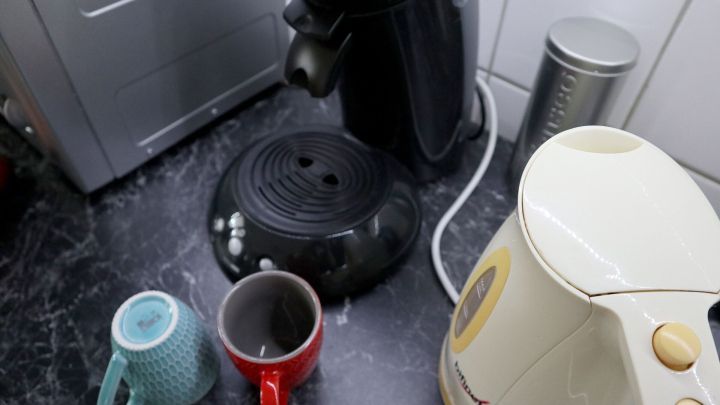 Kaffeekocher und Tassen stehen auf einer Arbeitsfl&auml;che in der Kaffeek&uuml;che, neben einem Wasserkocher und einer Kaffeedose.