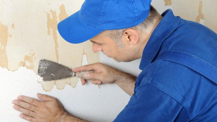 Ein Mann in blauer Kappe entfernt Tapete mit Spachtel von der Wand.