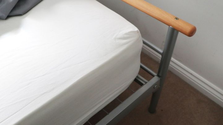 Ein Bett mit einem wei&szlig;en Matratzenbezug steht auf einem Teppich. Seine Holzleiste wirkt etwas abgenutzt.