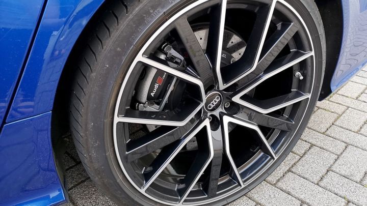 Ein Audi-Autoreifen mit gl&auml;nzender Felge steht auf grauem Pflasterstein, zeigt klare Details der Bremsanlage und Felge.