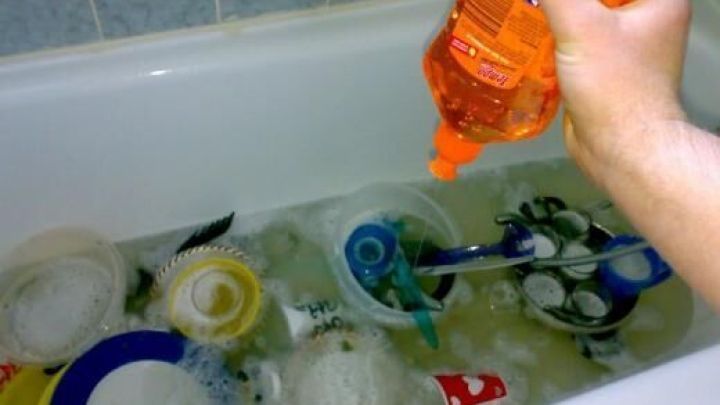 Verschiedene eingeweichte Geschirrteile schwimmen in einer Badewanne, w&auml;hrend jemand Sp&uuml;lmittel hinzugibt.