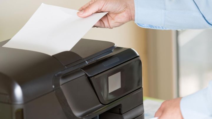 Eine Hand legt ein Blatt Papier in einen Drucker, w&auml;hrend der Drucker in einem hellen Raum steht.