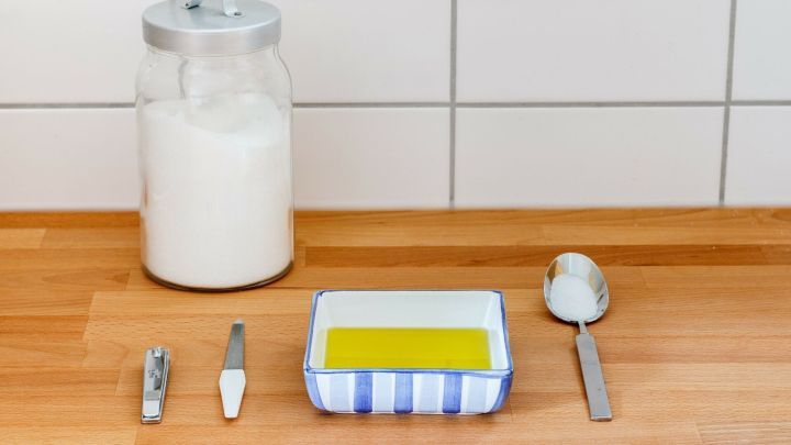 Ein Sch&auml;lchen mit &Ouml;l steht neben Zucker auf einem Holztisch, w&auml;hrend Werkzeuge bereitliegen, um die Handpflege vorzubereiten.
