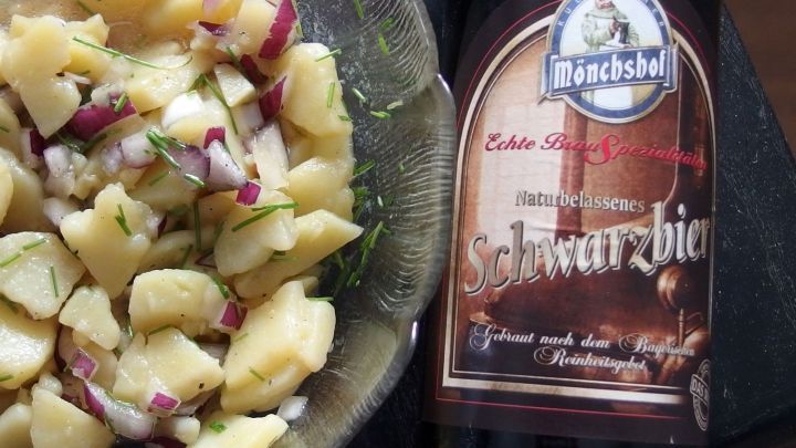 Ein Teller mit selbstgemachtem Kartoffelsalat und einer Flasche Schwarzbier daneben, alles auf einem Holztisch arrangiert.