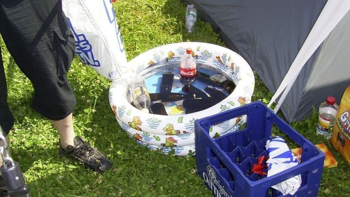 Flaschen schwimmen im Wasser eines kleinen Planschbeckens auf Gras neben einem Zelt. Eine blaue Kiste steht daneben.