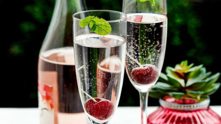 Zwei elegante Gl&auml;ser mit sprudelndem Sekt und einer Kirsche stehen neben einer Flasche Ros&eacute;wein auf einem Tisch.