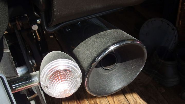 Die Auspuffanlage eines Motorrads steht im Fokus, w&auml;hrend sie in der Sonne gl&auml;nzt und im Hintergrund eine Holzfl&auml;che sichtbar ist.