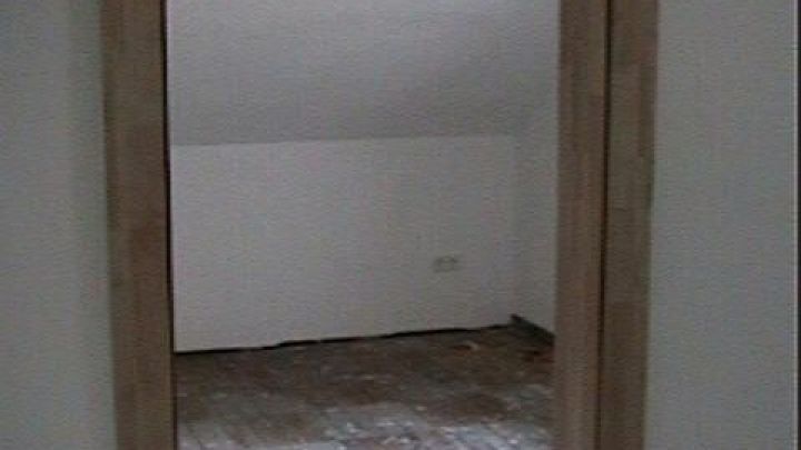 Holzt&uuml;rrahmen umrahmt leeren, unrenovierten Raum mit abgenutztem Holzboden und freiliegender Gl&uuml;hbirne an der Decke.