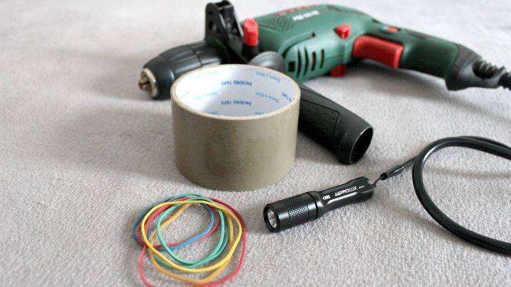 Eine Bohrmaschine liegt zusammen mit Klebeband, einem Taschenlampen und Gummib&auml;ndern auf einem hellen Untergrund.