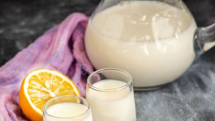 Ein klarer Krug mit einem wei&szlig;en Cocktail aus Wodka und Milch steht neben zwei kleinen Gl&auml;sern auf einem lila Tuch, mit einer Zitrone im Hintergrund.