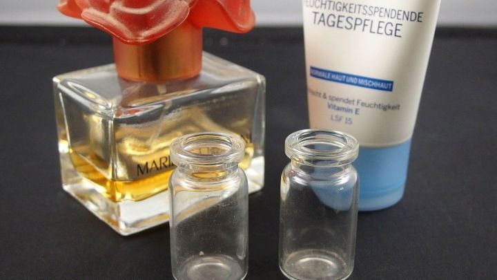 Eine elegante Parf&uuml;mflasche mit einem Blumenverschluss steht neben zwei kleinen, leeren Fl&auml;schchen und einer Tube Creme.