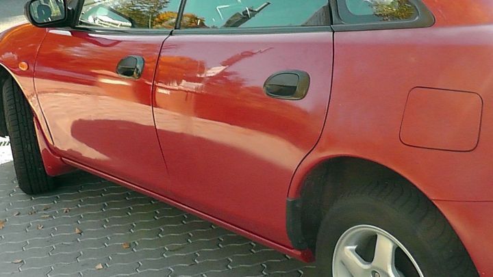 Ein rotes Auto parkt auf einem gepflasterten Parkplatz, m&ouml;glicherweise abbl&auml;tternde Stelle zeigt Anzeichen von Verrostung.