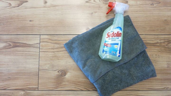Eine Flasche Sidolin Fensterreiniger liegt auf einem grauen Mikrofaser-Handtuch auf einem Holzlaminatboden.