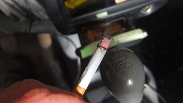 Eine Hand h&auml;lt eine brennende Zigarette, w&auml;hrend sie sich im Auto befindet, wo der Schaltkn&uuml;ppel sichtbar ist.