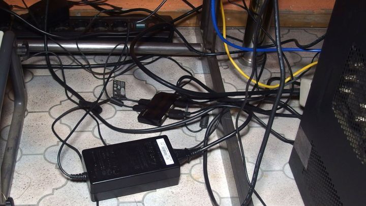 Ein unordentliches Durcheinander von Kabeln liegt unter einem Schreibtisch, w&auml;hrend ein Netzteil in der Mitte zu sehen ist.