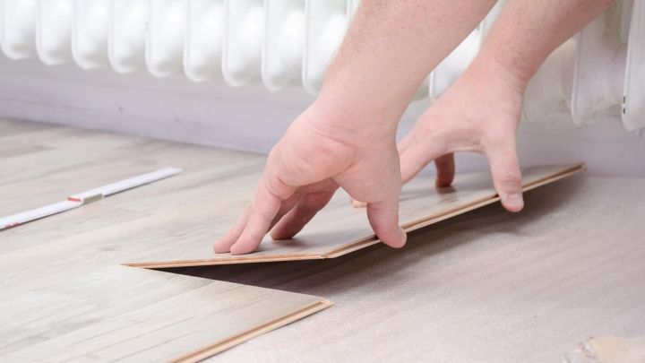 H&auml;nde legen ein Klicklaminat-Element auf den Boden, w&auml;hrend im Hintergrund ein Heizk&ouml;rper zu sehen ist.