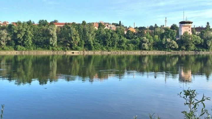 Ein ruhiger Fluss spiegelt den Himmel wider, umgeben von &uuml;ppigem Gr&uuml;n und einem Geb&auml;ude am Ufer im Hintergrund.