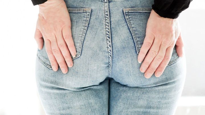 Eine Person h&auml;lt sch&uuml;chtern ihre H&auml;nde am Ges&auml;&szlig;, w&auml;hrend sie in l&auml;ssigen Jeans steht, m&ouml;glicherweise in einem hellen Raum.