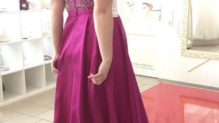 Eine Frau tr&auml;gt ein elegantes, rosa Kleid und steht in einem hellen Raum mit Spiegeln und einer roten Fl&auml;che.