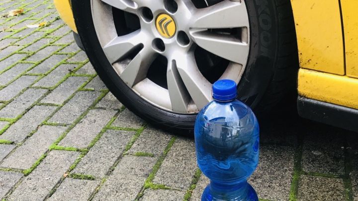 Eine blaue PET-Flasche steht am Bordstein neben einem gelben Auto, das auf einem gepflasterten Weg parkt.