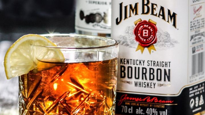 Ein Glas mit whiskeyhaltigem Getr&auml;nk und einer Limettenscheibe steht auf einem Holztisch, umgeben von Flaschen.