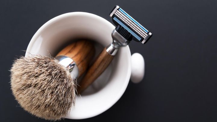 Ein Rasierer und ein Rasierpinsel liegen in einer eleganten Sch&uuml;ssel aus hellem Keramik auf einem schwarzen Hintergrund.