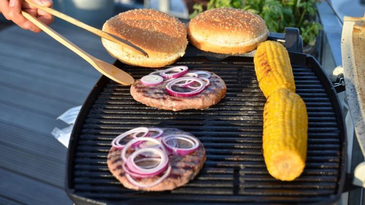 Saftige Burgerpatties grillen auf dem Balkon, w&auml;hrend frische Br&ouml;tchen und Maiskolben bereitliegen.