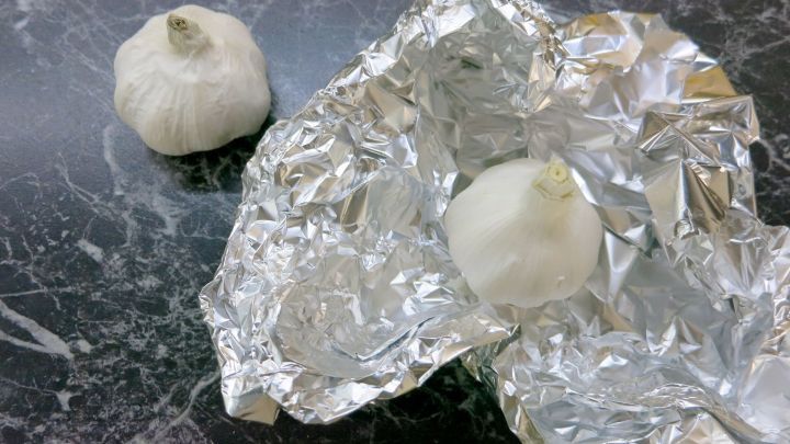 Eine Knoblauchzehe liegt auf einem Tisch, w&auml;hrend eine weitere in Alufolie eingewickelt gebacken wird.