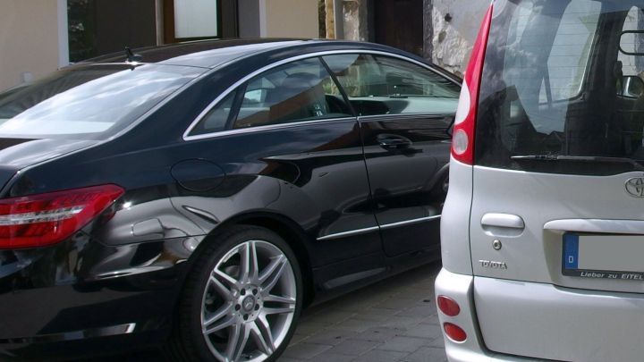 Ein schwarzes Auto mit Kratzern in der Lackierung steht neben einem hellen Fahrzeug auf einem Parkplatz.