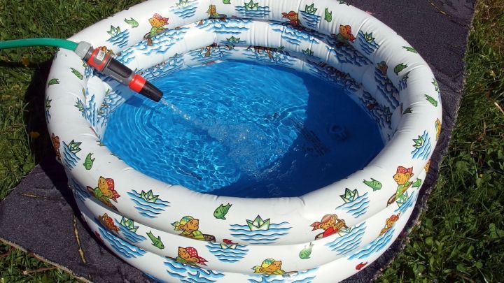 Ein buntes, aufblasbares Planschbecken wird mit einem Schlauch gef&uuml;llt, platziert auf einem alten Teppich im Garten.