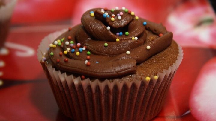 Ein Muffin mit Schokoladencreme und bunten Streuseln sitzt auf einem dekorativen Hintergrund mit roten Motiven.