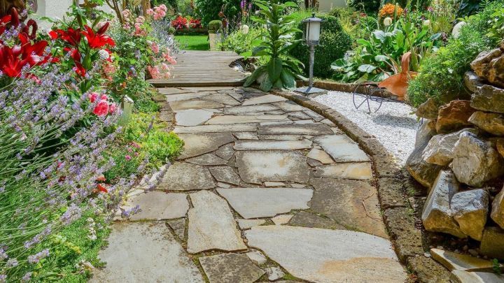 Ein gepflasterter Gartenweg schl&auml;ngelt sich durch eine bl&uuml;hende Landschaft voller bunter Blumen und Pflanzen.