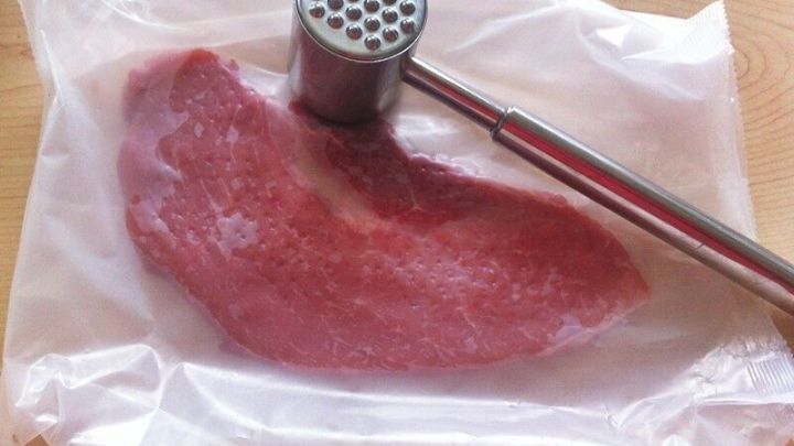 Ein Schnitzel liegt auf einem Blatt Plastikfolie, w&auml;hrend ein Fleischklopfer darauf schl&auml;gt, um es zart zu machen.