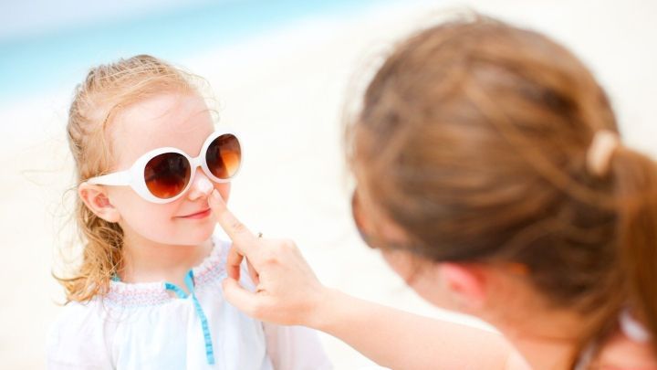 Ein M&auml;dchen mit Sonnenbrille sitzt am Strand, w&auml;hrend eine andere Person ihr sanft Sonnencreme auftr&auml;gt.
