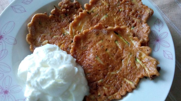 Zucchini-Puffer liegen goldbraun gebraten neben einem Klecks Sahne auf einem wei&szlig;en Teller mit Blumenmuster auf einer Tischdecke.