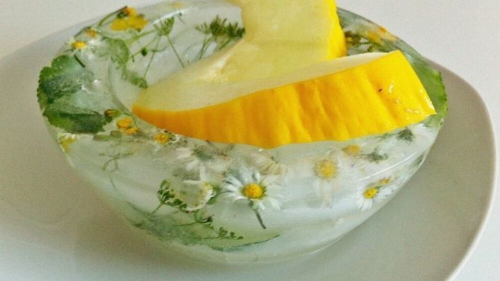 Eine dekorative Schale aus Eis zeigt eine runde Eisschale mit Blumen und einem Fruchtst&uuml;ck auf einem wei&szlig;en Teller.