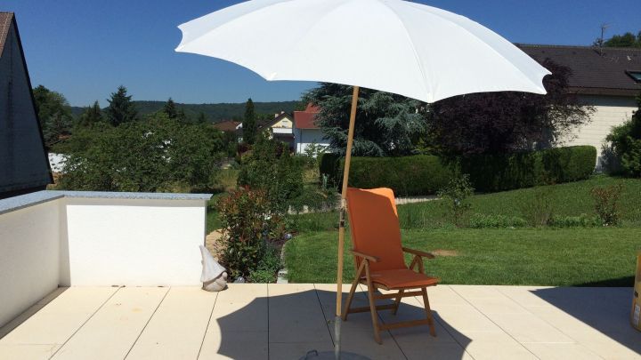 Ein Sonnenschirm steht auf einer Terrasse, w&auml;hrend er Schatten auf einen Liegestuhl wirft; dar&uuml;ber blauer Himmel.